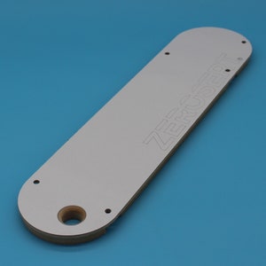 May include: White rectangular router template with the word "ZERO INSERT" printed on it. The template has four holes and a large hole in the center.
