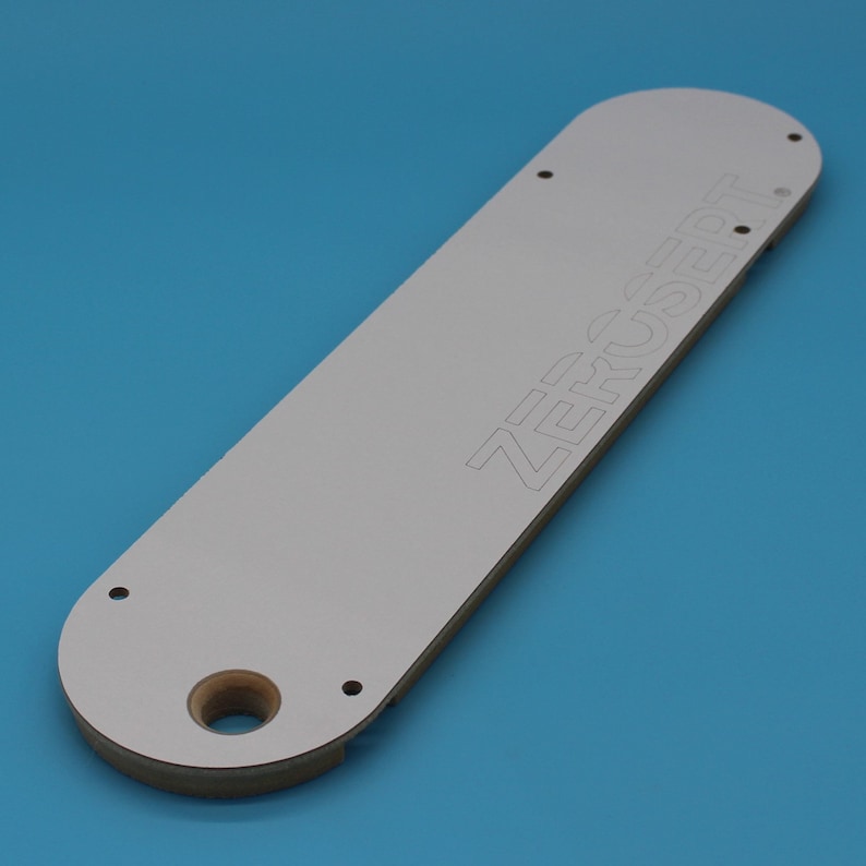 May include: White rectangular router template with the word "ZERO INSERT" printed on it. The template has four holes and a large hole in the center.