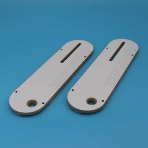 May include: Two white wooden router templates with the text "ZERUSERT" printed on them. The templates have a slot for a router bit and a hole for a dust collection port.
