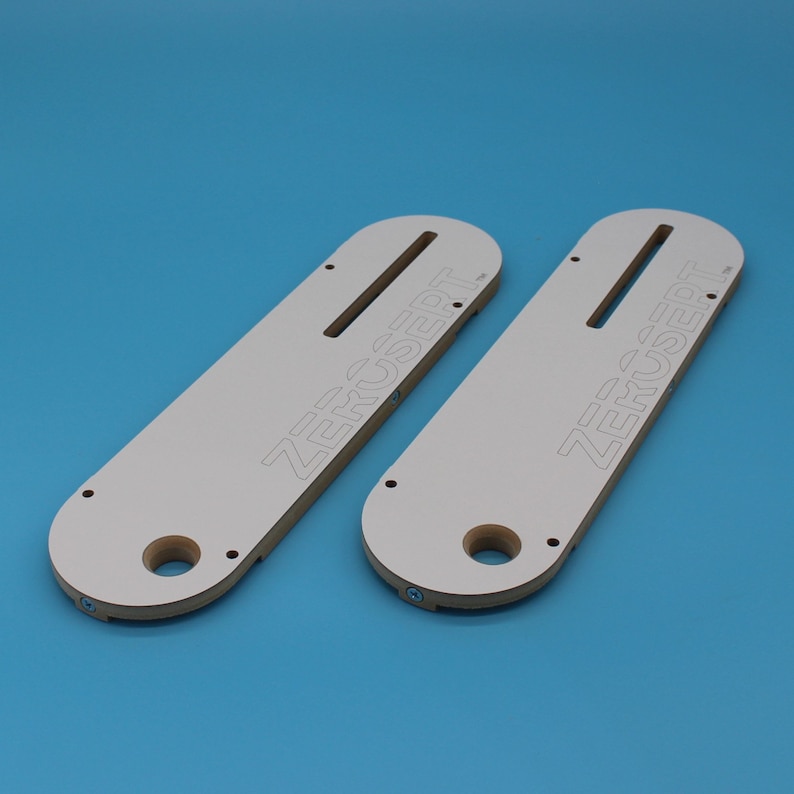 May include: Two white wooden router templates with the text "ZERUSERT" printed on them. The templates have a slot for a router bit and a hole for a dust collection port.