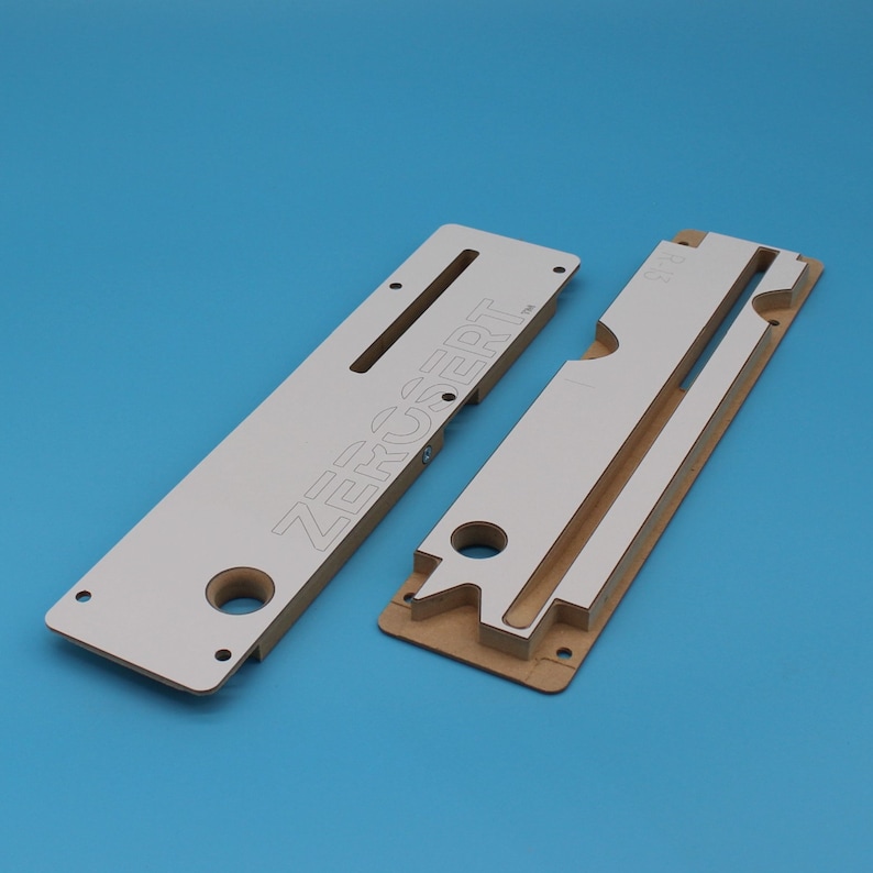 May include: Two white and brown wooden templates for woodworking projects. The larger template has the word "ZERUSERT" printed on it. The smaller template has a cutout for a hinge.