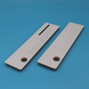 Pu&ograve; includere: Due mensole in legno bianco con la scritta "ZERCSERT" stampata su di esse. Le mensole hanno un foro al centro e una fessura nella parte superiore.