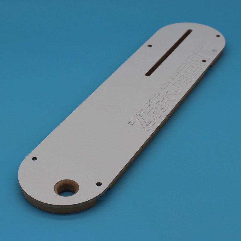 May include: White router table insert with the text "ZERO-CLEAR" printed on it. The insert has a slot for a router bit and several holes for mounting.