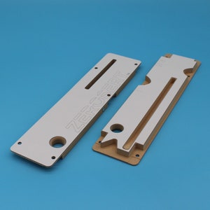 May include: Two white wooden templates for woodworking projects. The larger template has the word "ZERCSERT" printed on it. The smaller template has a cutout for a drawer slide.
