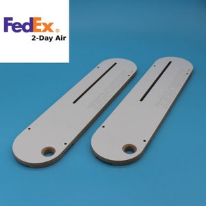 May include: Two white Zero Clearance inserts for a table saw, with a long, narrow slot and the brand name etched in the surface. The inserts have a rounded end with a circular hole. The background is a solid blue.