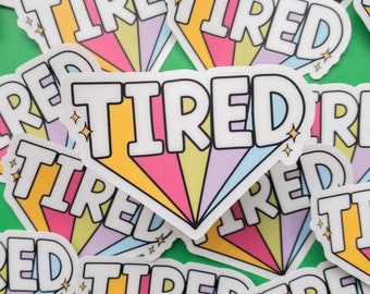 May include: A close-up of colorful stickers with the word "TIRED" printed in a rainbow font. The stickers are arranged in a random pattern on a green background.