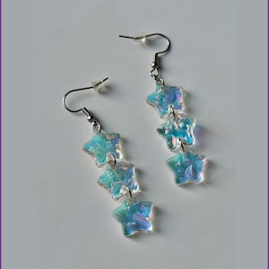 May include: A pair of dangling earrings featuring three iridescent star-shaped charms. The charms are suspended from silver-colored hooks. The earrings have a light blue and purple color scheme. The background is white with a rainbow stripe.