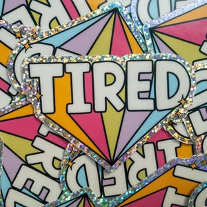 May include: A stack of colourful stickers with a rainbow design and the word "TIRED" in white letters. The stickers have a glittery finish.