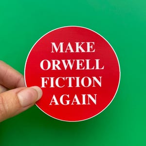 Puede incluir: Una pegatina redonda con fondo rojo y texto blanco que dice "MAKE ORWELL FICTION AGAIN". La pegatina se sostiene sobre un fondo verde.