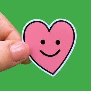 May include: A pink heart-shaped sticker with a smiling face. The sticker is being held by a finger.