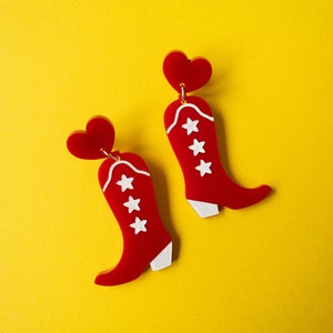 May include: A pair of red cowboy boot earrings with white star and trim details, and a red heart-shaped top. The earrings are set against a bright yellow background.