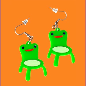 Pode incluir: Um par de brincos verdes em forma de cadeira, com ganchos prateados. Cada brinco apresenta um design de cadeira de desenho animado com um assento verde claro, olhos azuis e um sorriso vermelho. O fundo é laranja com uma faixa de arco-íris.