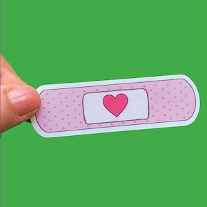 May include: A pink plaster with a white centre and a pink heart in the centre. The plaster is being held by a hand.