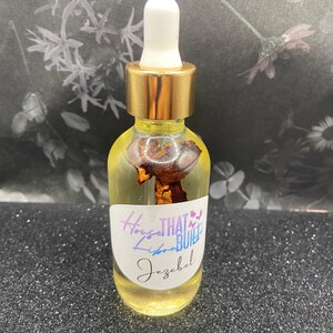 May include: A clear glass bottle with a gold cap and white dropper, filled with yellow liquid and a dried botanical element. The label reads "House That Built Libra Jezebel" in a script font. The background is a floral design.