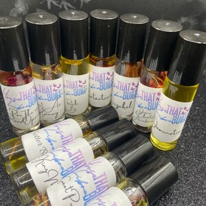 May include: A collection of roll-on essential oil bottles with black caps and labels that read "THAT BUILT." The bottles contain various colored oils and floral elements. The labels feature handwritten text, including "Better Being," "Sweet Pussy," and "Domination."