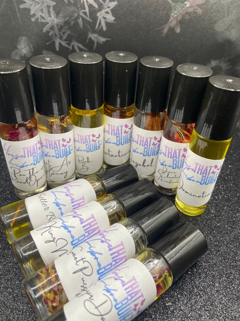 May include: A collection of roll-on essential oil bottles with black caps and labels that read "THAT BUILT." The bottles contain various colored oils and floral elements. The labels feature handwritten text, including "Better Being," "Sweet Pussy," and "Domination."