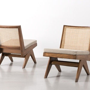 Op de afbeelding: Twee houten fauteuils met geweven rieten rugleuningen en lichtgekleurde kussens. De stoelen hebben een modern design uit het midden van de eeuw, met schuine poten en een laag profiel. Het hout heeft een warme, natuurlijke afwerking.