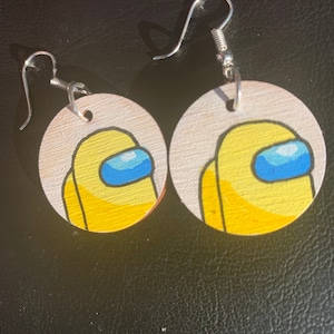 May include: A pair of wooden earrings with a yellow and blue Among Us character painted on each earring. The earrings are attached to silver hooks.