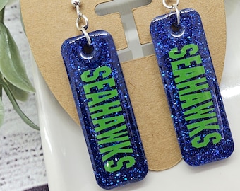 May include: A pair of blue glitter earrings with green text that says "Seahawks".