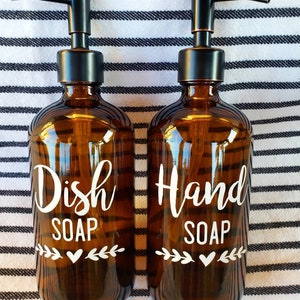 May include: Two amber glass bottles with black pump tops and white labels. The bottle on the left says "Dish Soap" and the bottle on the right says "Hand Soap". Both bottles have a small white heart and leafy design on the label.