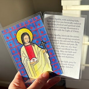 May include: A laminated prayer card featuring a colorful illustration of Saint Agatha holding a cross and a heart. The text on the card reads "Saint Agatha, with undying faith, hope, and love you heroically endured agonizing physical and spiritual trials and by your courage illuminated a dark world with the Light of Christ."