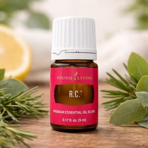 May include: A small brown glass bottle of Young Living R.C. Premium Essential Oil Blend. The bottle has a white cap and a pink label with the text "R.C." and "0.17 fl. oz. (5 ml)". The background includes green leaves and a lemon slice.