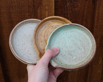 Pode incluir: Três descansos de colher de cerâmica feitos à mão em creme, castanho e verde menta. Cada um tem um design circular com uma pequena aba e um entalhe para apoiar os utensílios. O acabamento salpicado adiciona um toque rústico a estes utensílios de cozinha.