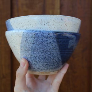 May include: Two ceramic bowls stacked, featuring a speckled design. The top bowl is light beige with dark speckles, while the bottom bowl has a blue and white speckled pattern. The bowls are handmade and suitable for serving food.