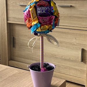 Puede incluir: Un ramo de chocolate hecho con una variedad de chocolates Cadbury Roses en un cubo morado con un palo a rayas rosa y amarillo y una cinta blanca con el texto "Feliz aniversario" en él.