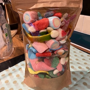 Puede incluir: Una bolsa de plástico transparente llena de una variedad de caramelos coloridos, incluyendo gusanos gomosos, malvaviscos y gominolas. La bolsa está sobre una superficie de lunares blancos y azules.