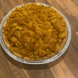 Puede incluir: Una tarta entera en una bandeja de aluminio plateado. La corteza de la tarta está rellena con un topping dorado y caramelizado hecho de copos de maíz. La tarta está sobre una superficie de madera marrón.