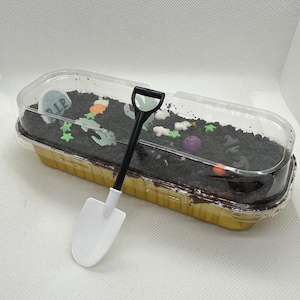 Puede incluir: Un recipiente de plástico transparente lleno de un pastel de tierra de chocolate decorado con dulces con temática de Halloween y una pequeña pala de plástico blanca. El pastel está cubierto con una lápida que dice "RIP".