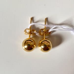Könnte beinhalten: Ein Paar goldene Kugelohrstecker. Jeder Ohrring hat eine kleine, polierte Kugel, die an einem Stecker mit einem Butterfly-Verschluss befestigt ist. Die Ohrringe liegen auf einer weißen Oberfläche, mit einer weißen Schnur.