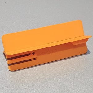 May include: An orange plastic phone stand with the word "POKER" printed on the side. The stand has a rectangular shape with a slot to hold a phone and two parallel lines on the front.