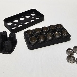 Puede incluir: Tres bandejas de plástico negro con 16, 12 y 12 depresiones circulares. Las bandejas están llenas de 12 discos de metal plateado con pequeños agujeros en el centro.