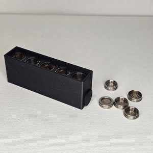 Puede incluir: Una caja de almacenamiento de plástico negro con seis compartimentos para guardar pequeños objetos metálicos redondos. La caja tiene un tapón de goma negro en un extremo. Seis objetos metálicos están dispersos sobre una superficie blanca.