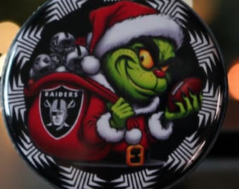 May include: A round, black and white decorative item featuring a green Grinch character in a Santa hat and suit, holding a football. The Grinch is carrying a sack with the Raiders logo. The item has a decorative border.
