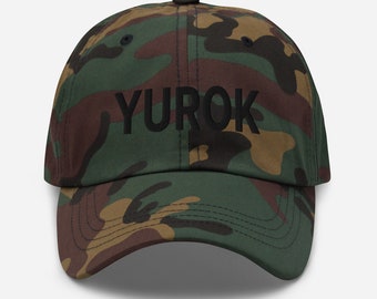 May include: A camouflage baseball cap with the word "YUROK" embroidered in black on the front.