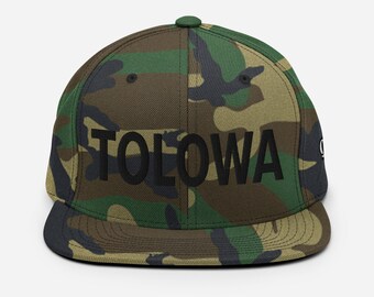 May include: A camouflage green and brown baseball cap with the word "TOLOWA" embroidered in black on the front.