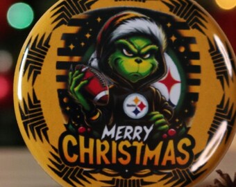 May include: A round, yellow Christmas ornament featuring a cartoon Grinch character wearing a Steelers hoodie and holding a football. The ornament has the words "MERRY CHRISTMAS" and the Steelers logo.