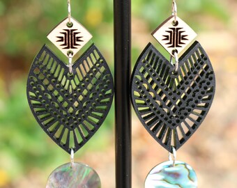 May include: Dangle earrings featuring a light-colored wooden diamond top with a black geometric design, a black laser-cut leaf-shaped middle piece with intricate patterns, and a round iridescent abalone shell charm at the bottom on silver hooks.