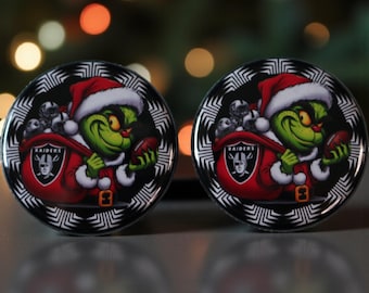 May include: Two round Christmas ornaments featuring the Grinch in a Santa hat, holding a football and a sack with Raiders logos. The ornaments have a black and white patterned border and are set against a blurred background.