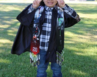 May include: A young person wearing a black graduation gown and cap, a plaid shirt, and blue jeans. They are holding up two fingers in a peace sign and have a colorful beaded graduation sash with the number 20 on it.