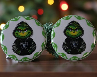 May include: Two round Christmas ornaments featuring a green Grinch character wearing a black Raiders hoodie. The Grinch has a mischievous grin and is set against a white background with a green geometric border. The ornaments are on a wooden surface.