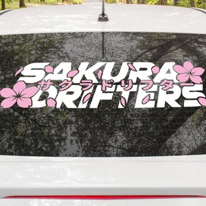 Peut inclure: Autocollant de fenêtre de voiture blanc avec l'inscription "SAKURA DRIFTERS" en lettres stylisées, accompagné de graphiques de fleurs de cerisier roses et de caractères japonais. L'autocollant est appliqué sur la vitre arrière d'une voiture blanche.