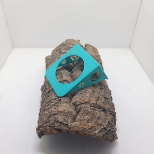 May include: A turquoise blue plastic 3D printed object with a circular cutout and a triangular shape. The object is resting on a piece of bark.