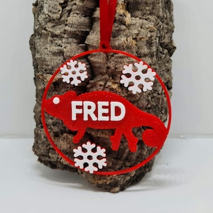 May include: Red Christmas bauble shaped like a chameleon with the name FRED printed in white. The bauble is decorated with white snowflakes.