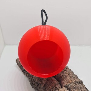 May include: A vibrant red, spherical bird feeder with a large opening and a black cord for hanging. The feeder is resting on a piece of textured, brown bark, set against a white background. The round shape and vibrant colour make it a striking garden accent.