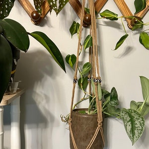 May include: A brown leather plant hanger with a macrame design and black and white beads. The hanger is holding a potted plant with green leaves.
