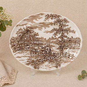 Könnte beinhalten: Eine weiße Keramik-Servierplatte mit braunem Transferdruck-Design. Die Szene zeigt eine Pferdekutsche vor einem Gebäude, mit Bäumen und einer Landschaft im Hintergrund. Die Platte hat einen gewellten Rand.